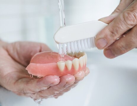 Close-up of hands cleaning a full upper denture with a denture brush under running water