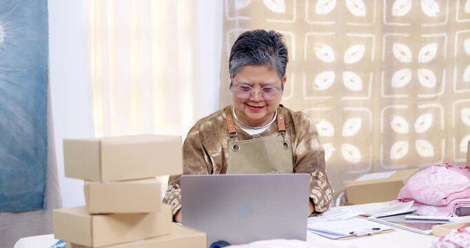 Senior asian woman entrepreneur smiles at laptop, confirms order checking, finishes invoice paperwork, stacks box for e-commerce parcel packing, small business shipping from home studio workshop.