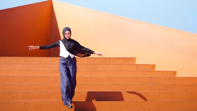 Carefree Asian Muslim Woman in Black Hijab Walking Down Orange Architectural Stairs with Arms Outstretched
