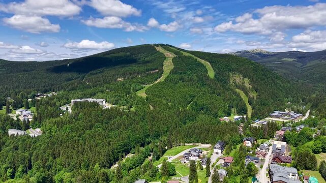 High oblique aerial view of forested mountain ridge with two wide grassy ski runs descending central slope, alpine village below, winding roads, rooftops, valley, and drifting cloud shadows across