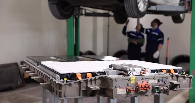 High-voltage lithium-ion battery pack displayed in the foreground while professional technicians service an electric vehicle in a modern automotive workshop.