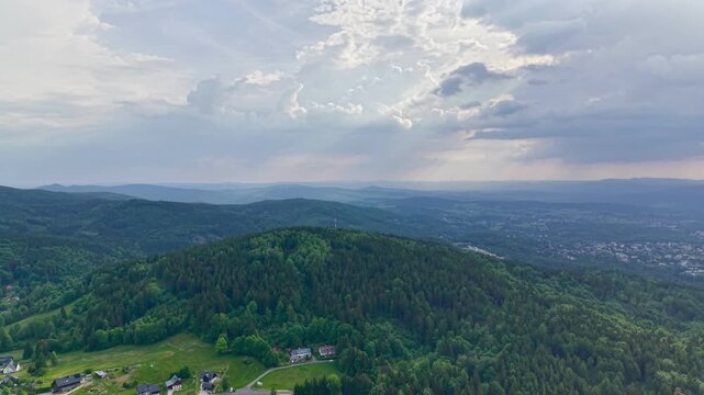 Aerial view of a tranquil rural countryside featuring rolling hills and scattered houses, captured from an elevated perspective. The scene is characterized by lush green vegetation.