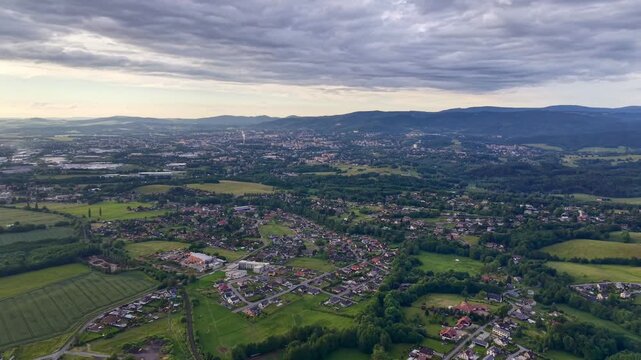 High oblique aerial view shows suburban neighborhood, detached houses, street grid, rectangular fields, woodland, and distant cityscape beneath layered overcast sky, with soft diffuse light and