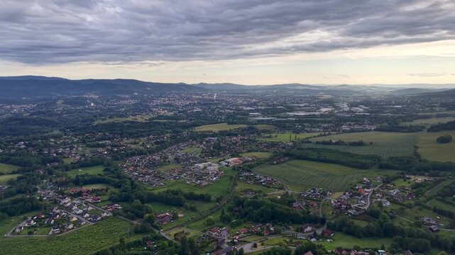 High oblique aerial view of valley town and surrounding countryside under heavy gray cloud cover, with soft diffuse light, low contrast, roads, rooftops, farmland, hills, and muted seasonal color