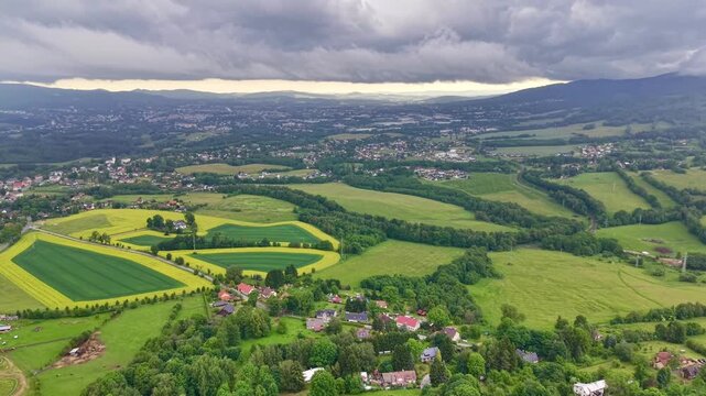 Wide oblique aerial view showing patchwork farmland, scattered village houses, winding country roads, green fields, and distant hills beneath heavy storm clouds, with bright horizon band adding