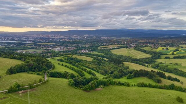 Sweeping rural landscape with layered hills, open pastures, mixed woodland, hedgerows, winding road, patchwork fields, bright crops, and soft horizon glow beneath dramatic cloud cover