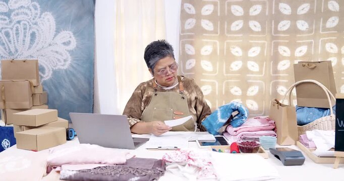 Older asian businesswoman pauses, reviews online order list, holds paperwork, checks inventory notes, box stack waits for pickup, shibori fabric packaging continues inside home studio workshop.