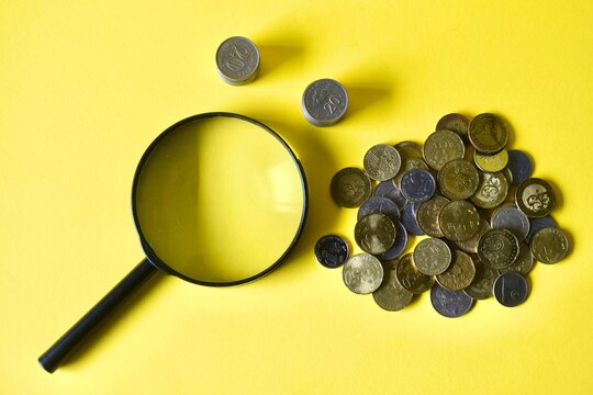 Top-down angle of a classic black magnifying glass placed next to arranged Malaysian coins and small coin stacks on a bright yellow backdrop. 