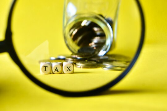A round magnifying glass frame focusing clearly on the word TAX spelled with wooden beads and scattered coins, set against an overturned glass jar on a yellow backdrop.