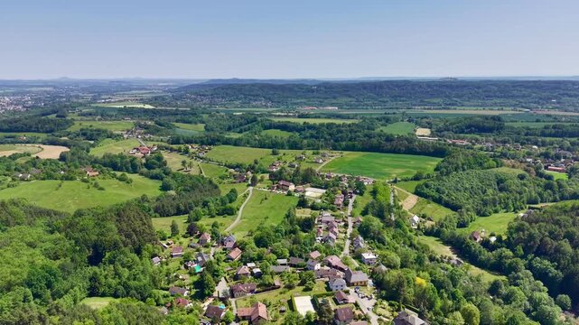 Drone view moves outward and slightly upward above clustered village roofs, garden plots, narrow roads, and scattered outbuildings, revealing patchwork fields, forested hills, and open blue sky