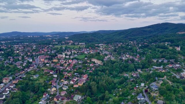 High aerial view of suburban homes, rooftops, streets, and green neighborhood edges beside forested hills beneath layered gray clouds, creating quiet rural landscape with broad scenic outlook