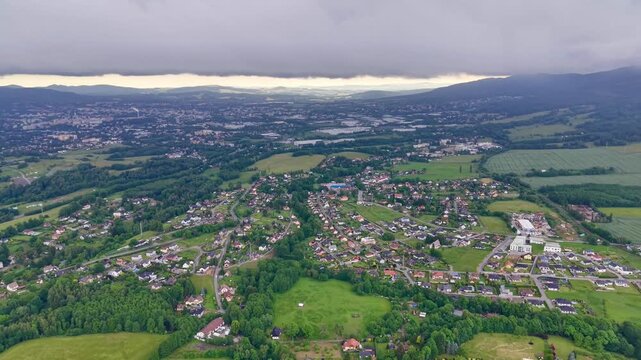 High oblique moving view shows suburban homes, streets, gardens, tree clusters, open fields, farmland, and distant town structures under layered cloud cover with soft shifting light across broad