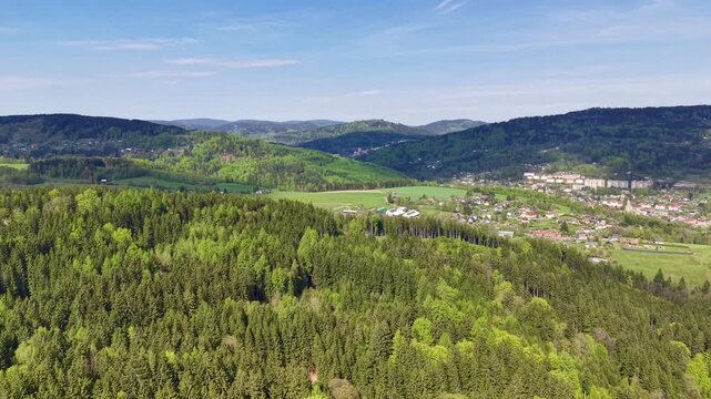 High viewpoint shows moving canopy of conifer and deciduous trees leading toward rural settlement, green fields, scattered houses, foothill slopes, and distant mountain ridges in calm daylight