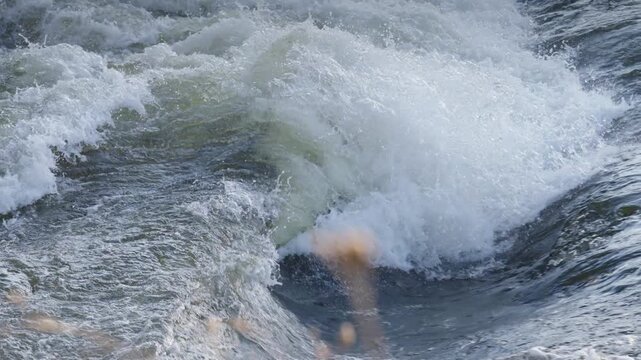 A powerful circular eddy swirls within fast-moving river currents, generating thick white foam. Glare from the spring sun reflects off the turbulent water surface.
