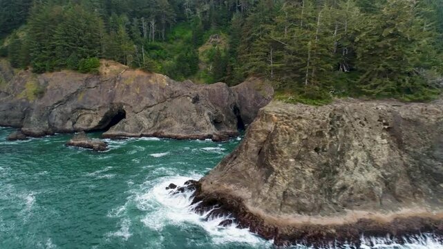 A cinematic, reversing wide-angle aerial drone shot of the rugged cliffs, unique rock formations, sea arches, and coastal forest at Samuel H. Boardman State Scenic Corridor on Oregon's southern coast.