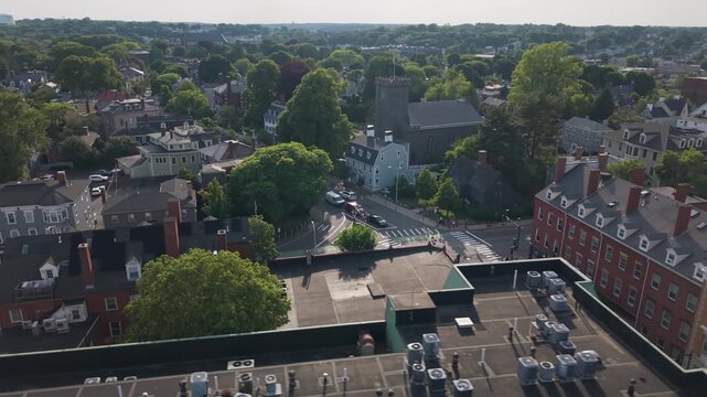 Salem Witch House anchors street corner in Salem Massachusetts, stone church tower rises above tree-lined street and parked cars below. Aerial drone flyover shot