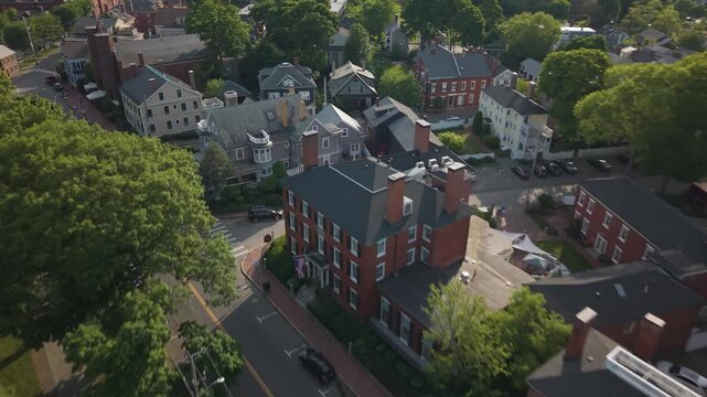 Historic three-story brick colonial mansion anchors tree-lined street corner in Salem Massachusetts, cars travel past federal-style homes and dense green summer trees. Aerial drone flyover