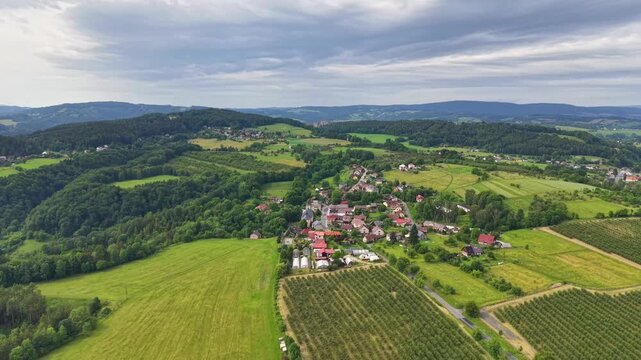 High aerial panorama shows rural village clustered across gentle hills beside patchwork farmland, winding roads, dense woodland, and wide horizon beneath soft overcast sky, with aerial, panorama