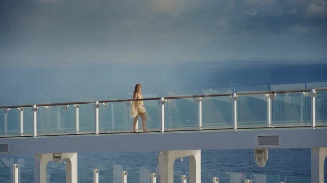 Young woman in summer dress walks along elevated glass walkway on cruise liner in Caribbean Sea. Wind blows her dress as she strolls barefoot on the modern ship deck with endless blue ocean horizon.