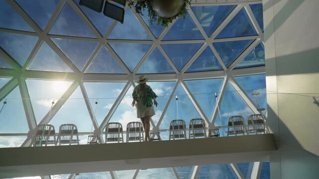 Stylish woman in white dress and straw hat strolls across elevated glass walkway aboard luxury cruise ship, geometric triangular skylights framing bright blue sky and fluffy clouds above ocean waters.