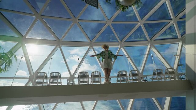 Woman stands with outstretched arms on glass balcony under geodesic dome ceiling aboard cruise liner in Caribbean Sea. Sunlight streams through triangular panels with blue sky and ocean views.