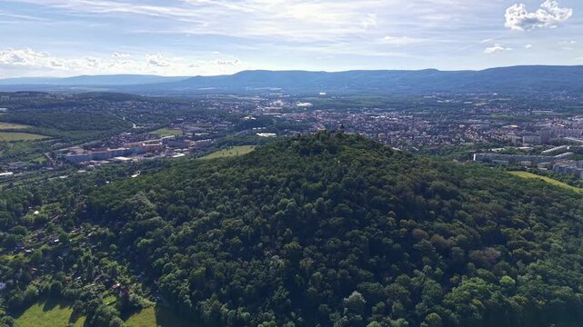 Elevated view reveals valley city nestled among forested hills with distant mountain ridges on horizon, broad skyline, green terrain, roads, rooftops, and open countryside under bright daylight