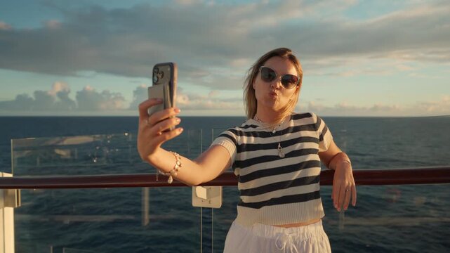 Young woman in striped shirt takes selfie with smartphone on cruise liner deck in Caribbean Sea. She makes kiss face while wind blows her hair against ocean horizon and dramatic sunset clouds.