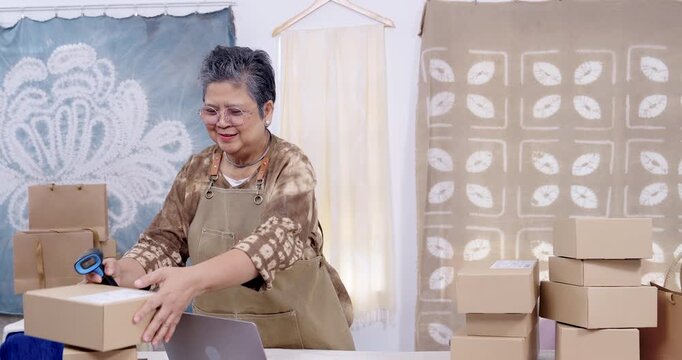 Senior asian retired craft seller scans parcel label with barcode reader, smiles, checks shipping status on laptop, organizes box stack for delivery workflow, slow motion fulfillment at home studio.