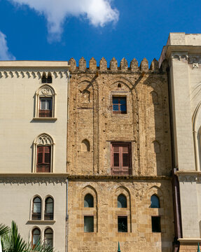 Fachada hist&oacute;rica de piedra antigua con arcos ciegos y almenas, contrastando con un edificio liso contiguo con ventanas ojivales bajo un cielo azul.