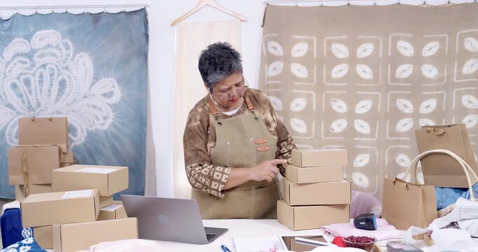 Senior asian entrepreneur stacks parcel boxes in slow motion, checks shipping label on carton, updates online order on laptop, keeps invoice and receipt ready, home studio dispatch continues.