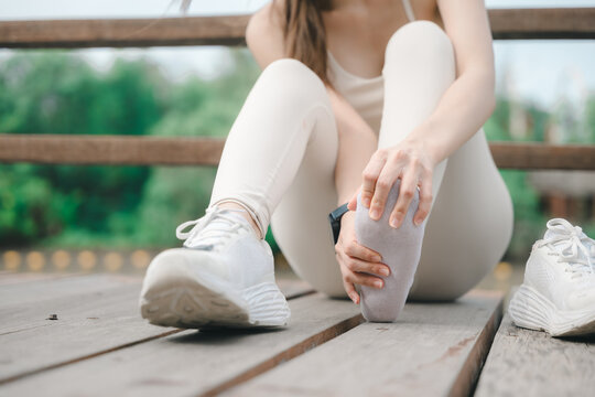 Sporty woman with pain in foot massaging sore sole after workout, sitting on wooden bridge with shoe removed suffering from sport injury or joint strain during outdoor fitness training