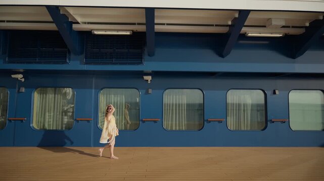 Young woman in summer dress walks along wooden deck of cruise liner in Caribbean Sea. Blue ship walls with large windows and curtains create nautical backdrop on sunny day.