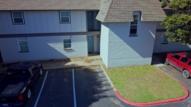 Daytime aerial shot of a multifamily housing building with adjacent parking lot, red curb, and tree-lined surroundings.