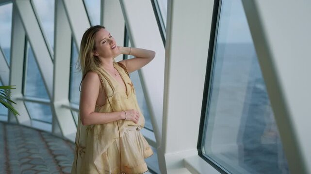 Young woman in elegant yellow dress stands by panoramic windows on cruise liner in Caribbean Sea gazing at ocean horizon. Modern ship interior with geometric glass design frames serene maritime scene.