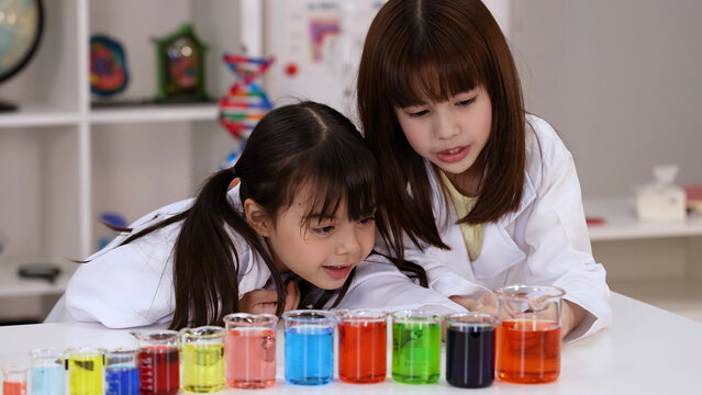 Two Elementary School Girls in Lab Coats Observing Colorful Chemistry Beakers