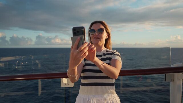 Young woman in striped shirt takes selfie photos on cruise liner deck in Caribbean Sea. She smiles and makes peace sign while holding smartphone against ocean horizon and dramatic sunset clouds.