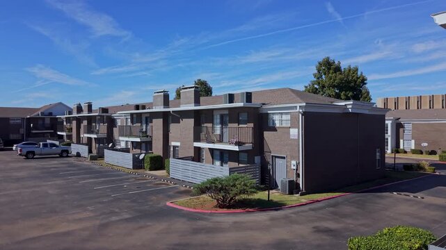 Sunny daytime shot of a brick multifamily housing complex with an empty parking lot, balconies, and fence.