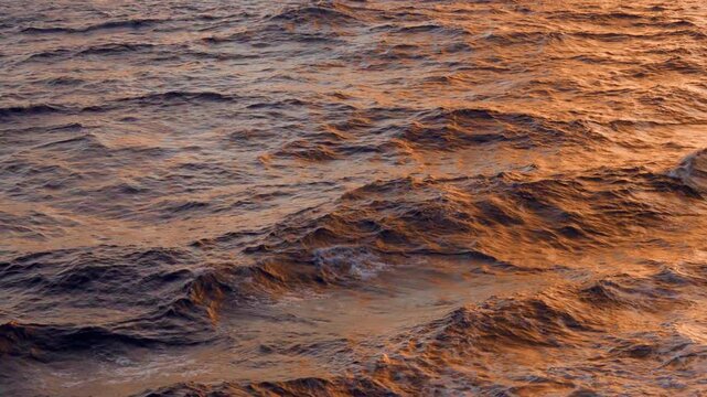 Choppy ocean waves illuminated by warm golden sunset light creating beautiful orange and copper reflections on the water surface. Rippling sea texture with dramatic evening colors.