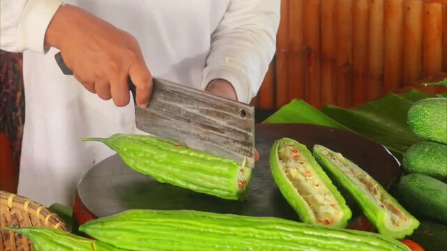 Chef slicing organic bitter gourd lengthwise on wooden board with cleaver 4K