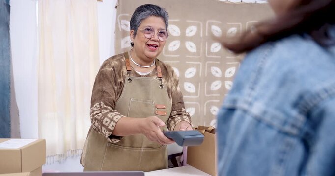 Retired woman small business owner laughs during checkout, holds card terminal, completes cashless payment, returns to online shop packaging and shipping label printing for artisan brand fulfillment.