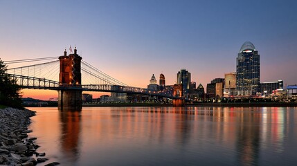 Fototapeta premium Iconic suspension bridge and modern city skyline illuminated at twilight with river reflections