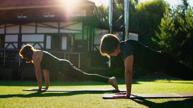 Elderly women practicing outdoor Pilates plank exercises together on yoga mats in slow motion, building strength, balance, and flexibility in a peaceful green setting