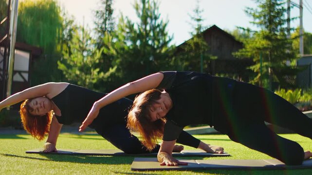 Elderly women performing outdoor Pilates side plank exercises together on yoga mats, moving in synchronized slow motion to improve balance, strength and flexibility