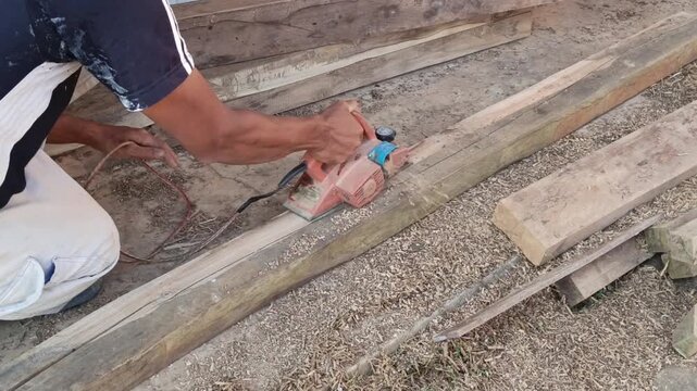Close-up stock video capturing a carpenter's hand pushing a power planer across a raw wood plank, removing rough layer shavings.