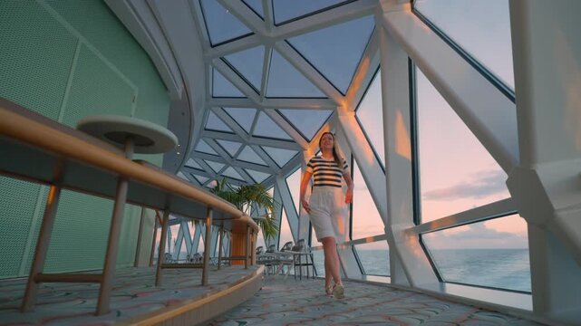 Young woman in striped shirt walks through luxurious cruise liner observation lounge with geometric glass windows overlooking the Caribbean Sea during golden hour sunset with pink sky reflections.