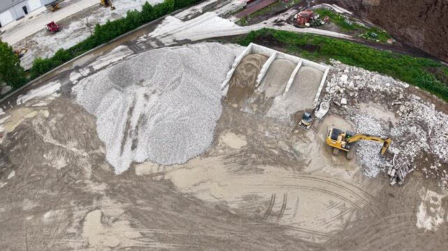 Drone view of a yellow loader working at the base of a towering pale aggregate stockpiles