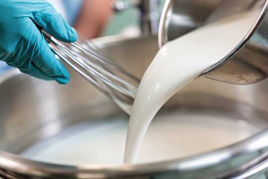 Gloved hand with whisk pours creamy liquid into stainless steel vat for food processing