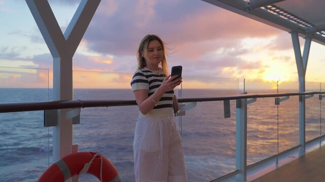 Young woman in striped shirt stands on cruise liner deck checking her phone during golden hour. Caribbean Sea stretches to horizon as dramatic sunset paints sky with purple and orange clouds.