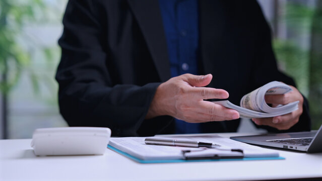 Male corporate accountant wearing dark suit counts paper currency dollar bills stack sitting behind office desk containing laptop computer calculator clipboard metal pen indoor.