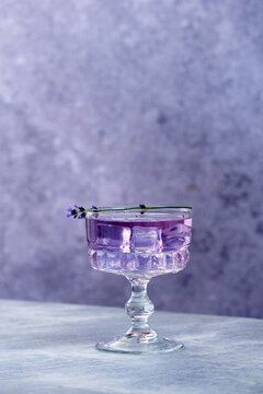 A glass filled with a purple drink sits on a light-colored table. Ice cubes fill the glass, and a sprig of lavender rests on the rim. The background is simple and plain.